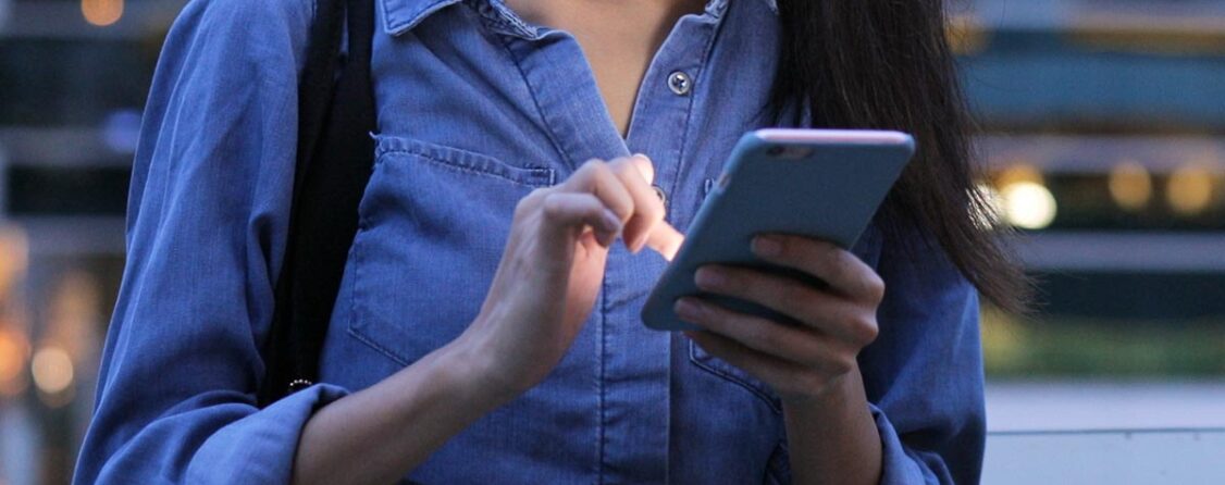 A photo of a woman with a backpack in a denim shirt typing on a smartphone.